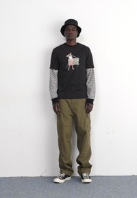 Camiseta gráfica negra de manga larga a rayas, pantalones cargo oliva y zapatillas negras. Lleva puesto un sombrero de cubo negro, de pie contra una pared blanca.