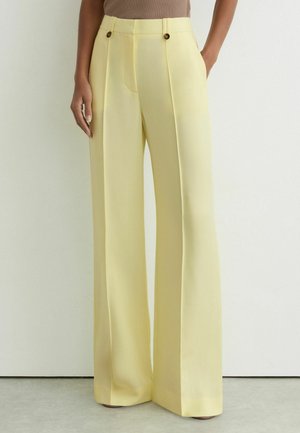Mujer con pantalones amarillos pálidos de talle alto y pierna ancha, con detalles de botones, de pie frente a una pared blanca lisa.