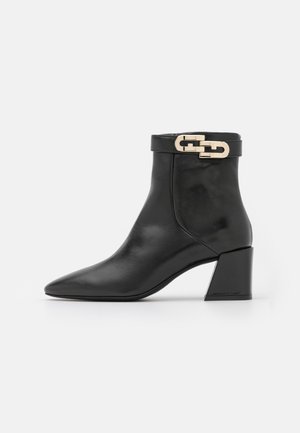 Bottine en cuir noir à bout pointu, talon bloc et détail d’une boucle chaîne dorée sur la bride latérale.