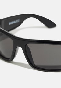 Lunettes de soleil noires avec une finition brillante, verres rectangulaires et monture épaisse ; elles présentent des verres teintés gris avec un design épuré.