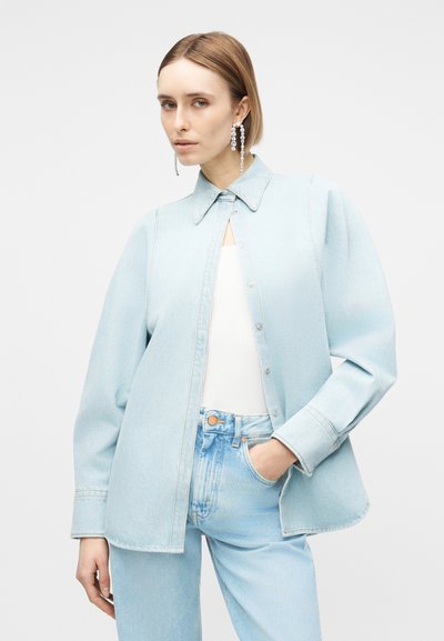 Femme aux cheveux courts portant une chemise en jean bleu clair surdimensionnée par-dessus un haut blanc, associée à un jean bleu clair et de longues boucles d'oreilles en cristal.