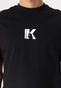 Zwarte katoenen T-shirt met een wit grafisch logo dat een profielsilhouet en de letters "IK" toont, geplaatst op de linkerkant van de borst.
