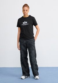 T-shirt noir avec le logo "ALPHA INDUSTRIES", associé à un pantalon noir ample avec des motifs floraux, et des baskets blanches.