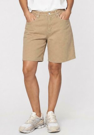 Persoon die beige knielange shorts met zakken, witte sneakers en een wit overhemd draagt, staand tegen een effen achtergrond.