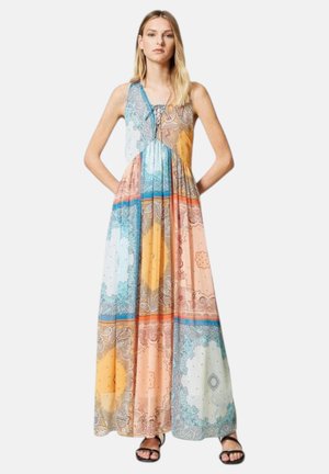 Donna che indossa un vestito maxi patchwork senza maniche multicolore con motivi paisley e sandali neri, in piedi con le mani in tasca.