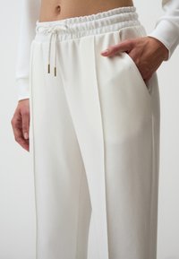Witte, elastische broek met een trekkoord en gouden hardwareaccenten, met zijzakken en een gladde, textuurovergetekende stof.