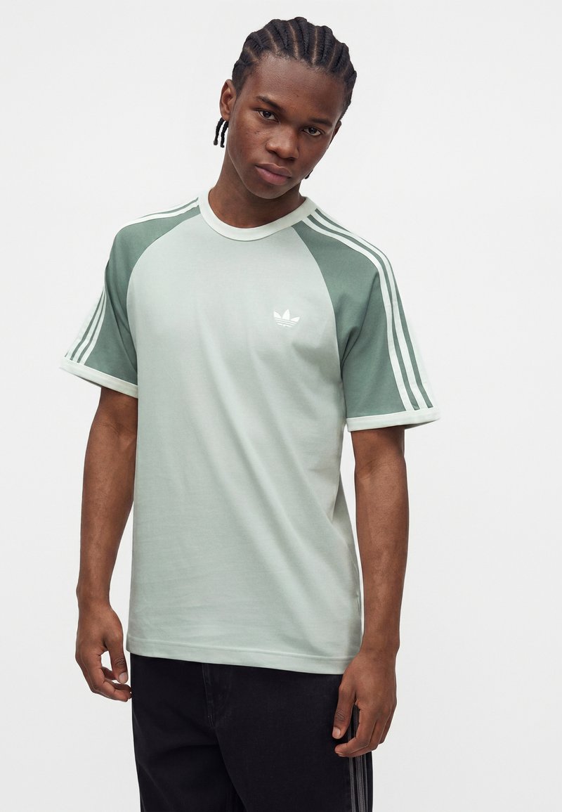Ung mand med flettet hår iført en lysegrøn Adidas t-shirt med hvide striber på skuldrene og sorte bukser, stående foran en ensfarvet baggrund.