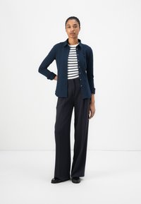 Camicia a maniche lunghe blu navy con bottoni sopra una maglietta bianca e blu a righe, abbinata a pantaloni a vita alta blu navy a gamba larga e scarpe basse nere.