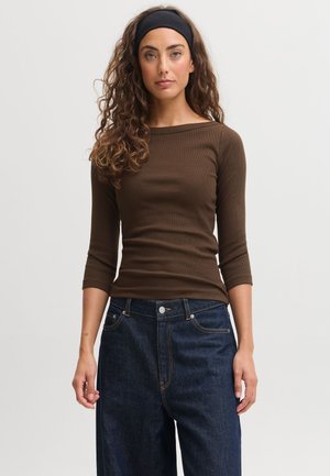 Femme aux cheveux bouclés portant un bandeau noir, un haut marron cintré à manches longues et un jean bleu foncé ample, debout devant un fond uni.