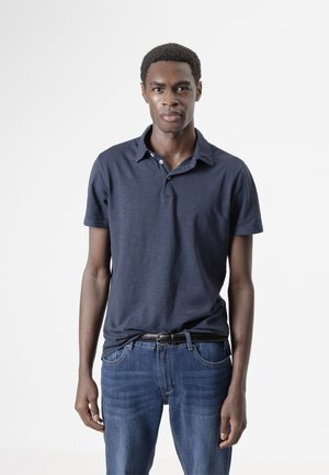 Joven con piel oscura y cabello corto, vestido con una camiseta polo azul marino y jeans azules, de pie sobre un fondo blanco liso.