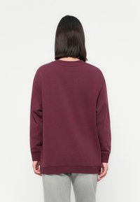 Personne aux cheveux foncés aux épaules portant un sweat-shirt bordeaux à manches longues et un pantalon gris clair, vue de dos.