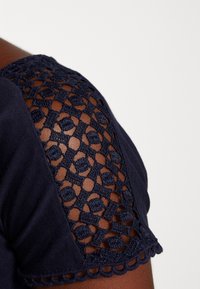 Granatowa koszulka z teksturowanym, szydełkowanym panelem na ramieniu i falbaniastymi brzegami. Gładki materiał i misterny wzór ażurowy podkreślają detale.