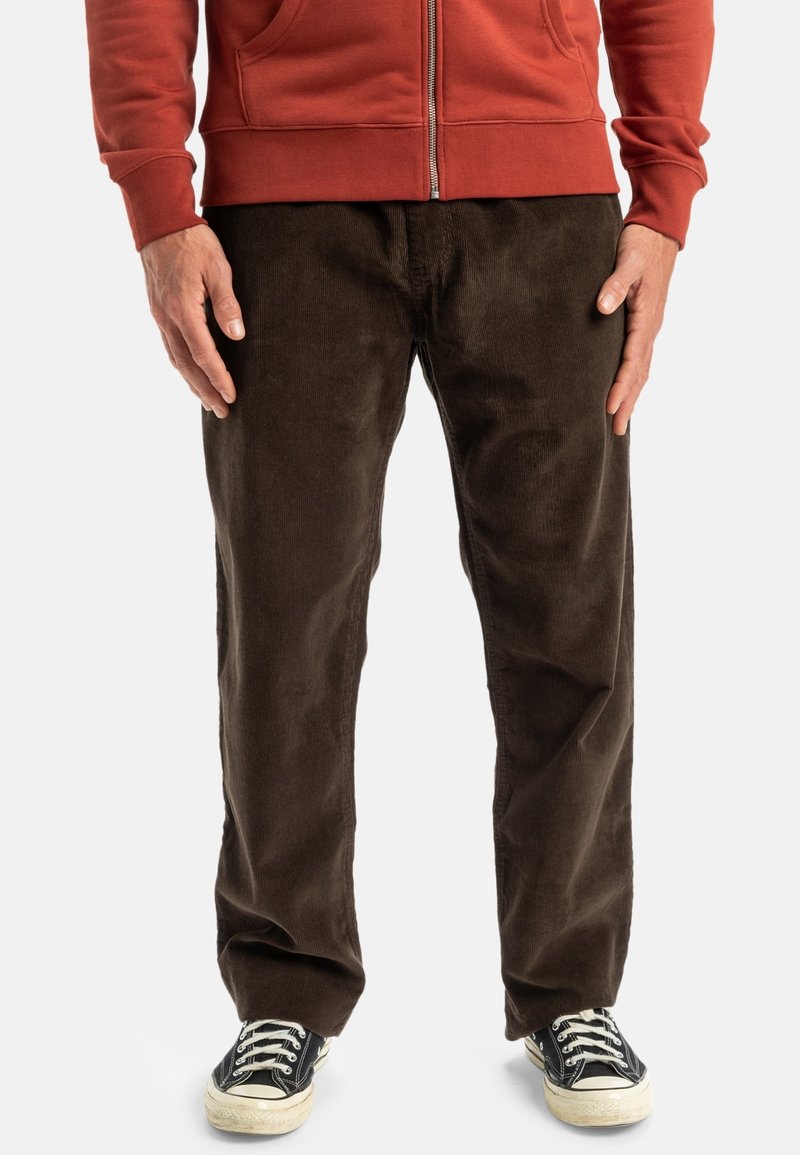 Homme portant un pantalon en velours côtelé marron foncé, des baskets noires et blanches, et un sweat à capuche zippé couleur rouille, debout avec les mains détendues le long du corps.