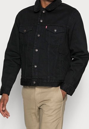Homme portant une veste noire à boutons avec col sherpa et pantalon beige, main dans la poche sur fond blanc.