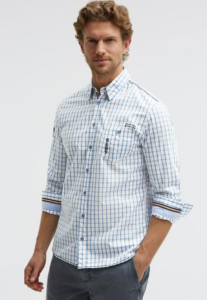 Mann, der ein weißes und hellblau kariertes Hemd mit aufgerollten Ärmeln und dunkle Hose trägt, mit einer Hand in der Tasche stehend.