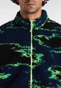 Fleece jack met een marineblauw en groen abstract patroon, hoge kraag en een contrasterende gele rits aan de voorkant. Zachte textuur.