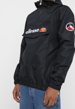 Personne portant un coupe-vent à capuche noir de la marque ellesse avec une grande poche avant et un écusson montagne sur la manche, associée à un jean bleu.