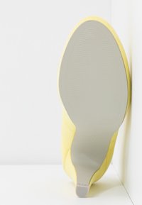 Chaussure à talons hauts en daim jaune avec une semelle en caoutchouc grise lisse présentant des motifs texturés. La chaussure a un design à bout pointu.