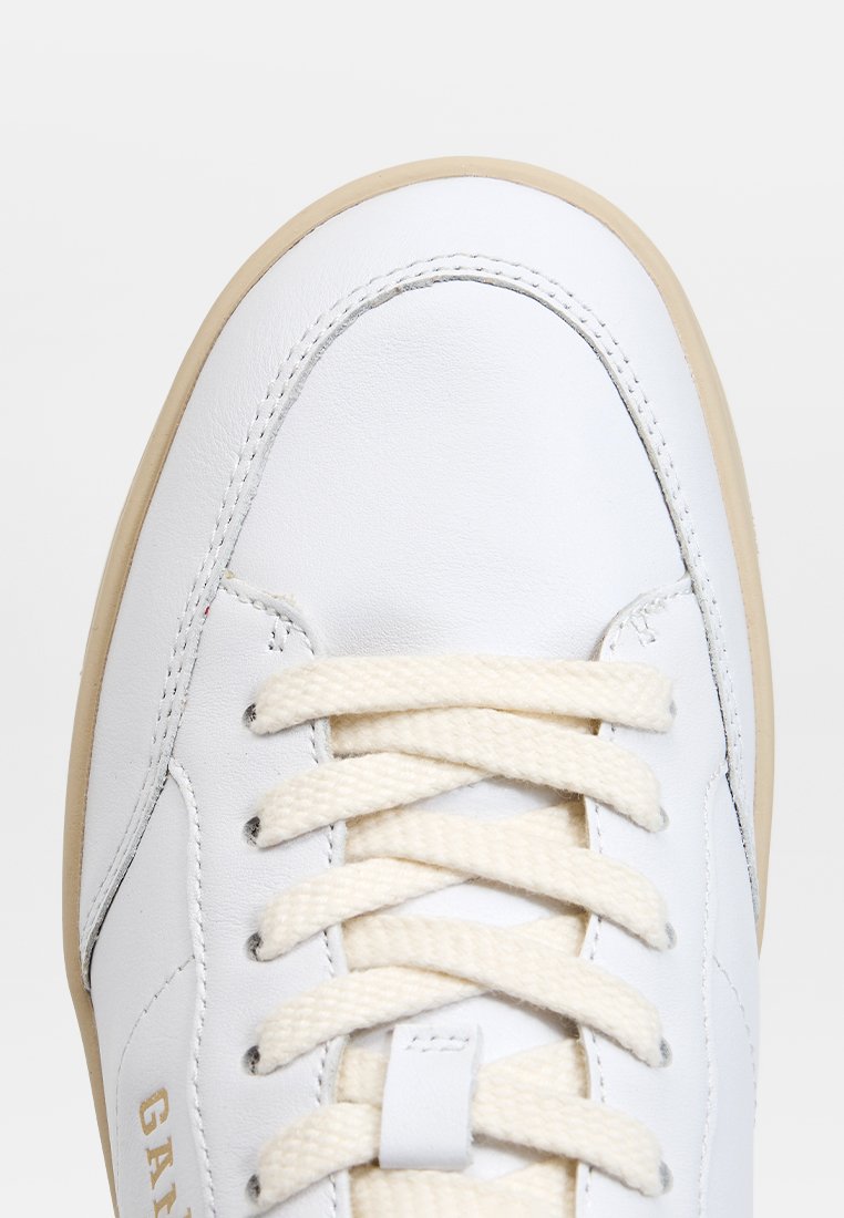 Zapatilla de piel blanca con puntera redonda, suela de goma beige, cordones crema y una sutil marca en el costado. Textura suave con detalles de costura.