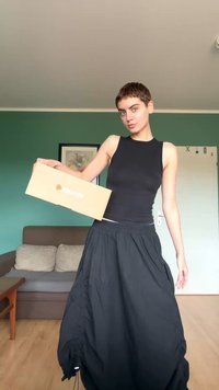 Personne aux cheveux courts portant un haut noir sans manches et une longue jupe noire tient une boîte en carton dans un salon aux murs verts.