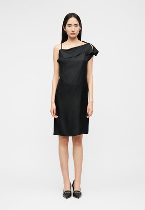 Donna che indossa un abito nero asimmetrico senza maniche con dettaglio a volant e tacchi neri a punta, in piedi davanti a uno sfondo chiaro e uniforme.