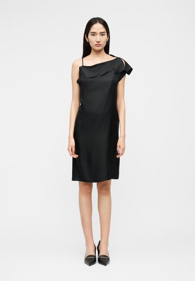 Femme portant une robe noire asymétrique sans manches avec un détail volanté et des escarpins noirs à bout pointu, debout devant un fond clair uni.