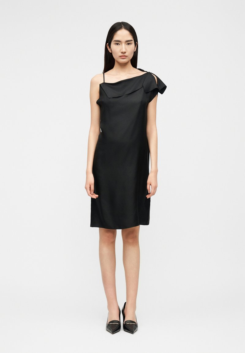 Femme portant une robe noire asymétrique sans manches avec un détail volanté et des escarpins noirs à bout pointu, debout devant un fond clair uni.