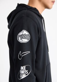 Melna kapuce ar priekšējo kabatu, ar baltiem logo grafikiem uz piedurknēm. Teksts ietver "Nike Sports Club" un stilizētu Nike logo.