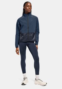 Marineblauer Windbreaker mit einem kontrastierenden dunkelblauen unteren Bereich, kombiniert mit marineblauen Leggings und weißen Sneakers mit schwarzen Akzenten.
