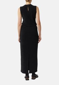 Kobieta w czarnej, bezrękawnikowej sukience midi z rozcięciem z tyłu i zapięciem na guziki, w połączeniu z czarnymi balerinami i przezroczystymi czarnymi rajstopami.