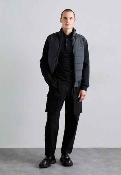 Black padded vest over a black polo, paired with loose black cargo pants featuring pockets. Model wears polished black shoes. Plain background.