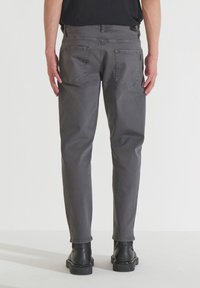 Los pantalones de mezclilla gris presentan un diseño de pierna recta con dos bolsillos traseros y una textura sutil. Usados con botas negras, se muestran desde la parte de atrás.