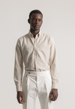 Hombre con camisa de botones beige y pantalones blancos de cintura alta, de pie contra un fondo liso claro con una expresión neutral.