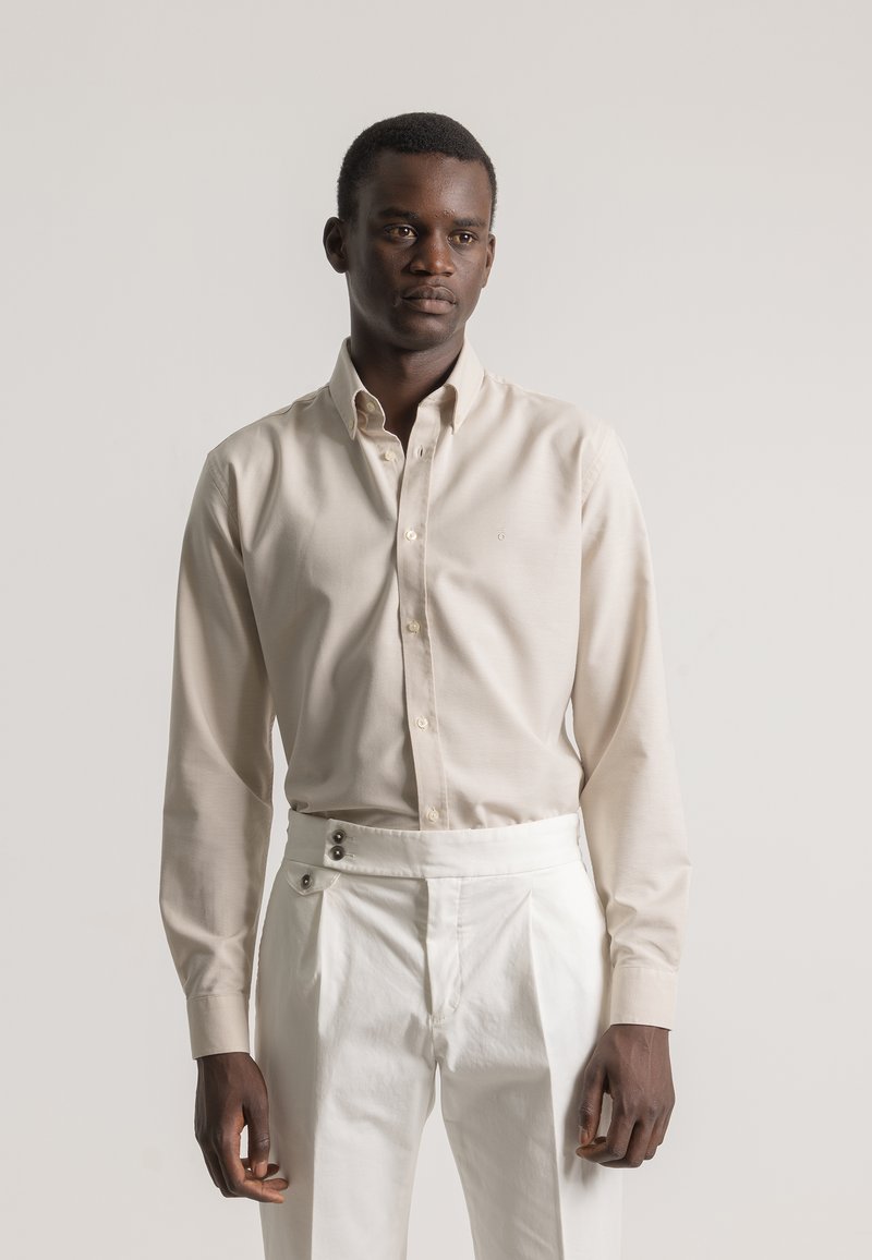 Hombre con camisa de botones beige y pantalones blancos de cintura alta, de pie contra un fondo liso claro con una expresión neutral.