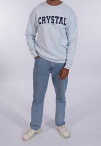 Felpa azzurra chiara con "CRYSTAL" in lettere blu navy in grassetto, abbinata a jeans azzurri chiari e sneakers bianche. Collo girocollo e maniche lunghe.