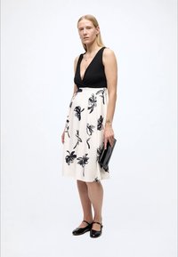 Top negru, fără mâneci, cu un decolteu adânc în V; fustă albă cu modele florale negre; asortate cu un plic negru și pantofi comozi.