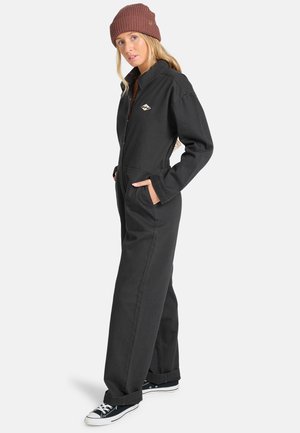 Jonge vrouw die een zwarte jumpsuit, een bruine gebreide muts en zwarte sneakers draagt, staand met handen in de zakken tegen een effen achtergrond.