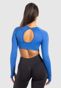 Donna che indossa un crop top blu a maniche lunghe con un'apertura circolare sul retro e leggings neri a vita alta, vista di spalle.