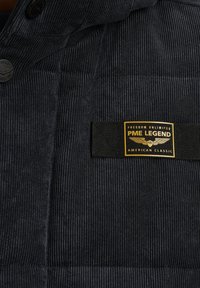 Veste en velours côtelé bleu foncé avec un design matelassé, quincaillerie noire et étiquette PME Legend de couleur or avec le texte "American Classic".
