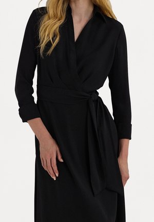 Femme portant une robe portefeuille noire à manches longues avec col et ceinture nouée à la taille, debout devant un fond clair uni.