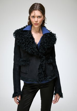 Femme portant une veste noire bouclée bordée de fourrure sur une chemise bleue à col et un pantalon noir, debout devant un fond clair uni.