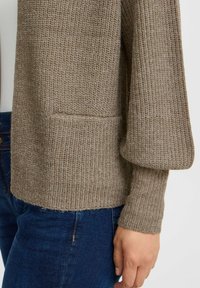 Cárdigan de punto en tonos beige-marrón, con un ajuste holgado, textura acanalada y un bolsillo en la parte frontal. Los extremos de las mangas son ligeramente acampanados.