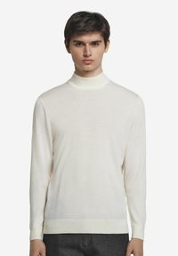 Lätt, krämfärgad polotröja i mjukt stickat material. Har lång ärmar och ribbade muddar, med en figurnära silhuett och minimal textur.