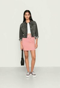 Jovem mulher vestindo uma jaqueta de denim preta, um top branco cropped, uma saia rosa, tênis estampados e segurando uma pequena bolsa preta contra um fundo simples.