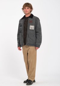 Jeune homme debout portant une veste gris foncé avec des écussons, un pull à motifs marron, un pantalon en velours côtelé beige et des chaussures noires sur fond blanc.
