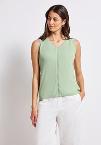 Top sans manches en V de couleur vert clair, réalisé en tissu lisse. Présente une couture centrale avec des détails de couture délicats. Associé à un pantalon blanc.