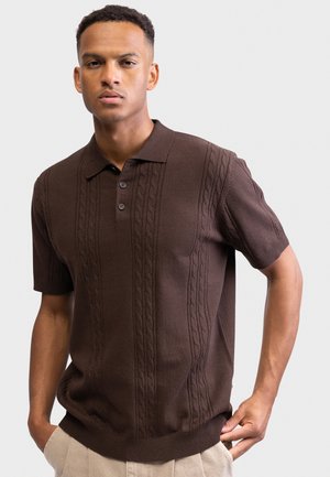 Fiatal férfi barna rövid ujjú kötött galléros pólóban, melyen kábelminták vannak, bézs nadrágban, az egyik kezét a zsebébe téve pózol fehér háttér előtt.