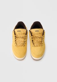 Dzeltenas zamšādas sporta kurpes ar brūniem akcentiem, kas ietver teksturētas paneles, apaļas šņorītes un baltu gumijas zoli. Uz mēles redzams Etnies logotips.