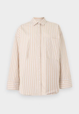 Jeune femme portant des lunettes rondes, une chemise beige rayée ample sur un haut sombre, un pantalon beige large et des chaussures marron, debout contre un mur gris uni.