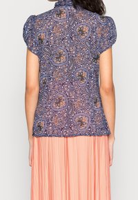 Blusa blu con motivo paisley, maniche a sbuffo, colletto alto e orlo leggermente svasato, indossata su una gonna rosa plissettata.
